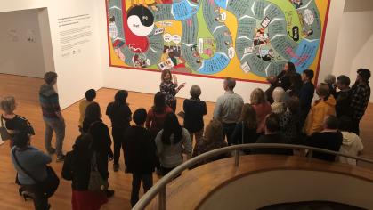 A group of people listens to a curator speak in front of a large image similar to a board game